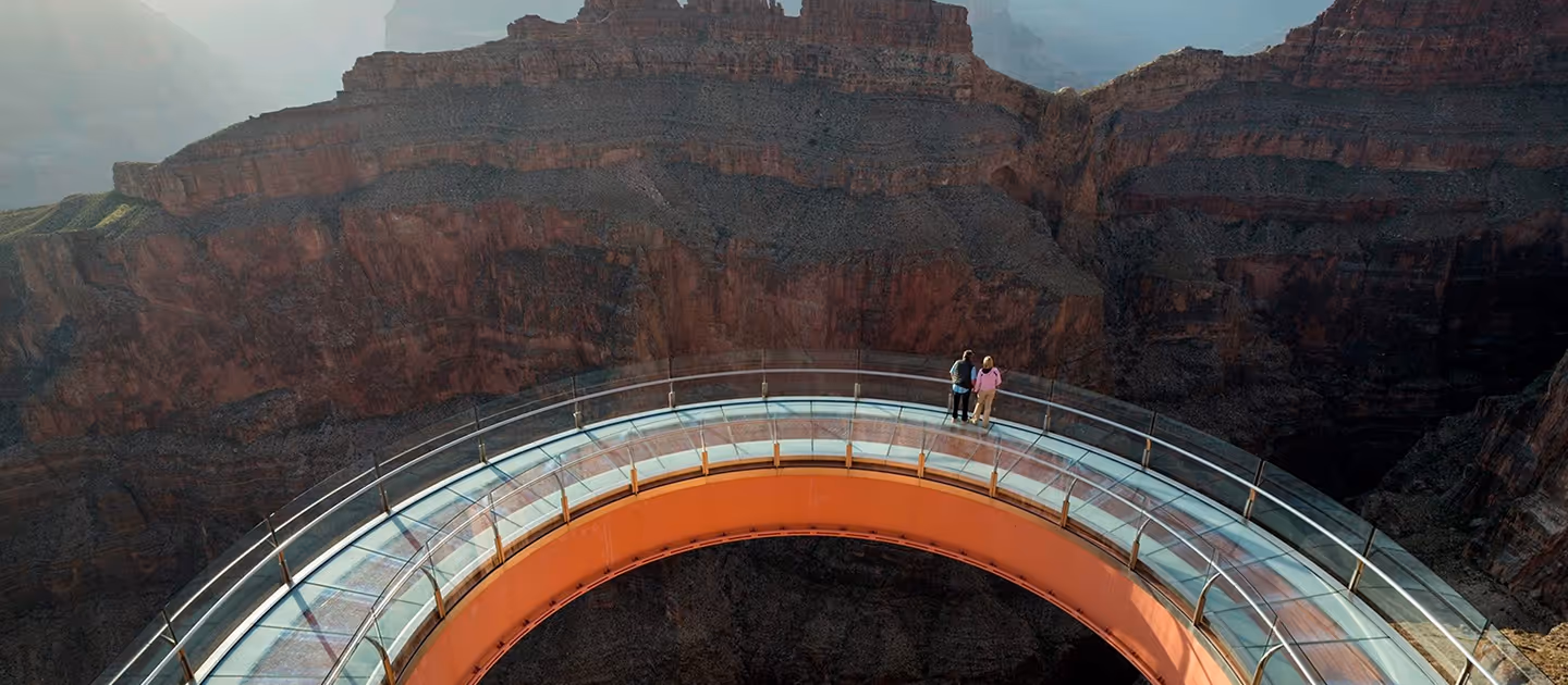 Two people standing on a curved glass observation deck overlooking the Grand Canyon cliffs.